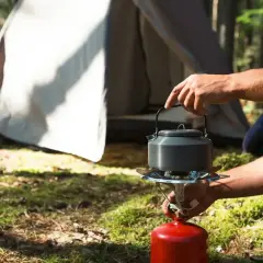 Ręka trzyma czajnik nad kuchenką turystyczną zasilaną z czerwonej butli gazowej. W tle namiot. Różne rodzaje butli gazowych są kluczowe dla biwakowania.