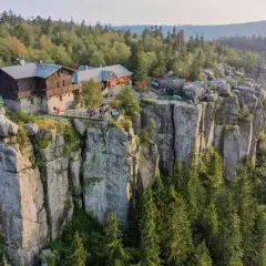 Schroniska PTTK na skalnym urwisku, otoczone lasem. Widać turystów odpoczywających na tarasach.