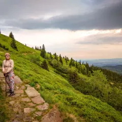 Karpacz-Śnieżka czas wejścia. Kobieta na kamiennym szlaku, zielone zbocza górskie, chmury na niebie.