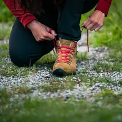 Przygotowanie do wędrówki: osoba klęczy, sznurując brązowe buty trekkingowe z czerwonymi sznurówkami. Zostawia 1,5 cm luzu w butach, by stopy miały komfort.