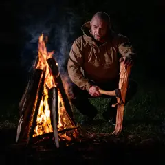 Mężczyzna z siekierą przygotowuje drewno do ogniska bez dymu. Płomienie tańczą w ciemności, tworząc ciepłą atmosferę.