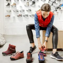 Kobieta przymierza buty trekkingowe, zastanawiając się, o ile powinny być większe. W tle widać inne buty i osobę w turkusowych spodniach.